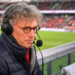 Peter Vandenbempt met headset aan een commentaarpositie in een voetbalstadion.