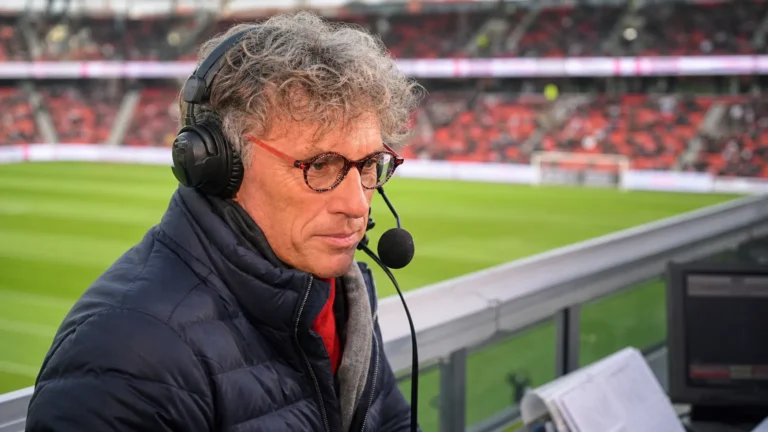 Peter Vandenbempt met headset aan een commentaarpositie in een voetbalstadion.