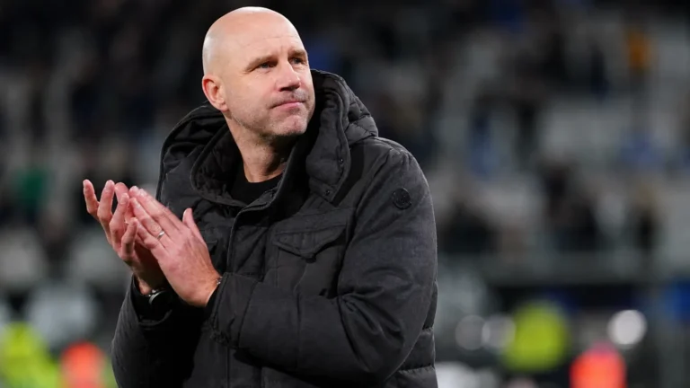 Een niet-spelende clubvertegenwoordiger in een donkere jas applaudisseert langs de zijlijn van een voetbalstadion.