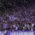 Beerschot-supporters in paars en wit vieren in een volle tribune met slierten.