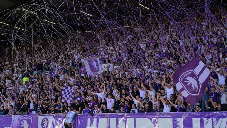 Beerschot-supporters in paars en wit vieren in een volle tribune met slierten.
