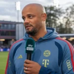 Een trainer in blauwe clubkleding spreekt tijdens een interview in een stadionomgeving.