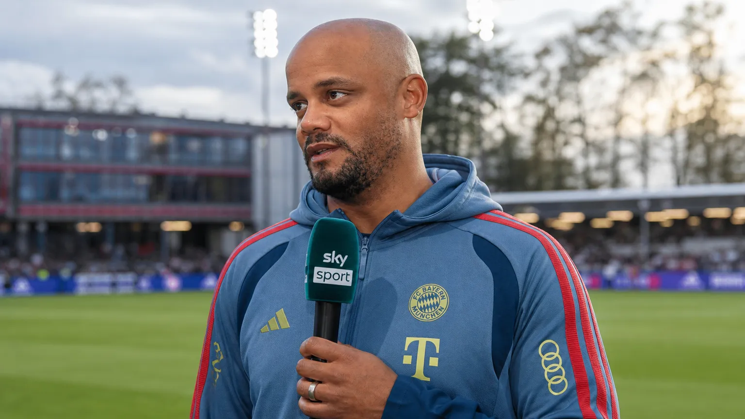 Een trainer in blauwe clubkleding spreekt tijdens een interview in een stadionomgeving.