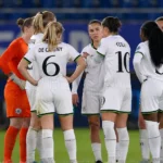 Speelsters in witte tenues vormen een hechte kring op het veld.