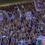 Een volle tribune met paars-witte voetbalsupporters, vlaggen en slingers.