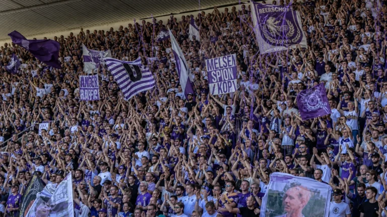 Een volle tribune met paars-witte voetbalsupporters, vlaggen en slingers.