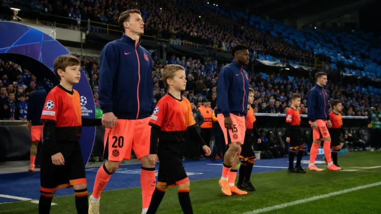 Voetballers lopen met jonge mascottes het veld op voor een avondwedstrijd.