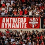 Een volle groep rood-wit geklede voetbalsupporters op een stadiontribune met spandoeken.
