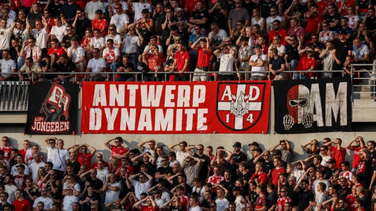 Een volle groep rood-wit geklede voetbalsupporters op een stadiontribune met spandoeken.