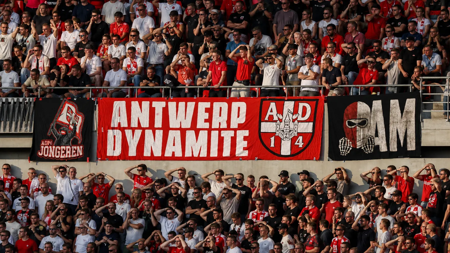 Een volle groep rood-wit geklede voetbalsupporters op een stadiontribune met spandoeken.