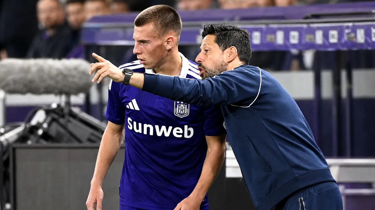 Een Anderlecht-trainer geeft aan de zijlijn aanwijzingen aan een speler in paars tenue.