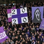 Een volle paars-witte voetbaltribune met supporters die grote spandoeken omhooghouden.