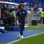 Malick Fofana in donkere trainingskleding langs de zijlijn van een voetbalveld.