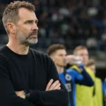 Een coach of staflid in donkere trui kijkt vanaf de zijlijn naar spelers.