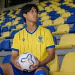 Kota Takai poseert met een bal in een geel STVV-shirt voor blauw-gele stadionzitjes.