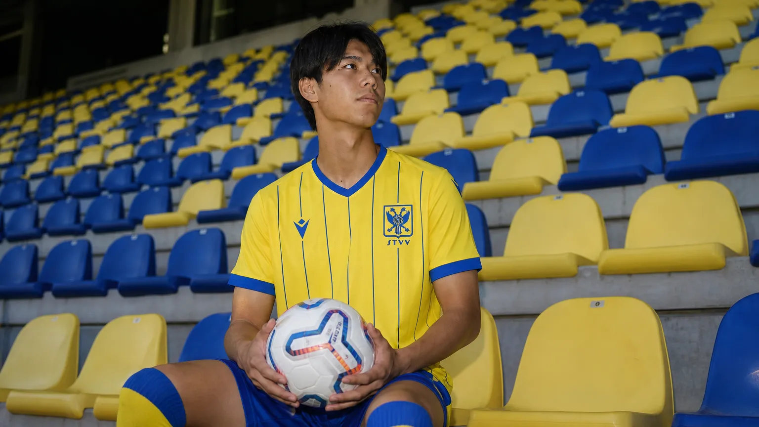 Kota Takai poseert met een bal in een geel STVV-shirt voor blauw-gele stadionzitjes.