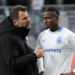 Een Club Brugge-coach spreekt langs het veld met een jonge speler in een wit Club Brugge-tenue.