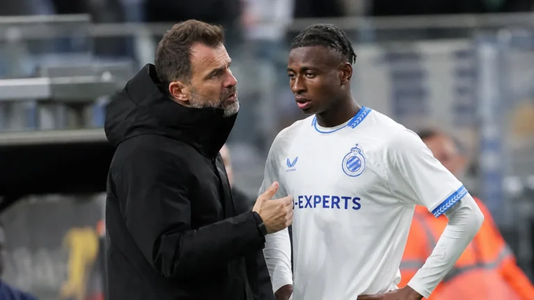 Een Club Brugge-coach spreekt langs het veld met een jonge speler in een wit Club Brugge-tenue.
