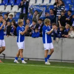Voetballers in blauw-witte shirts groeten supporters langs de rand van het veld.