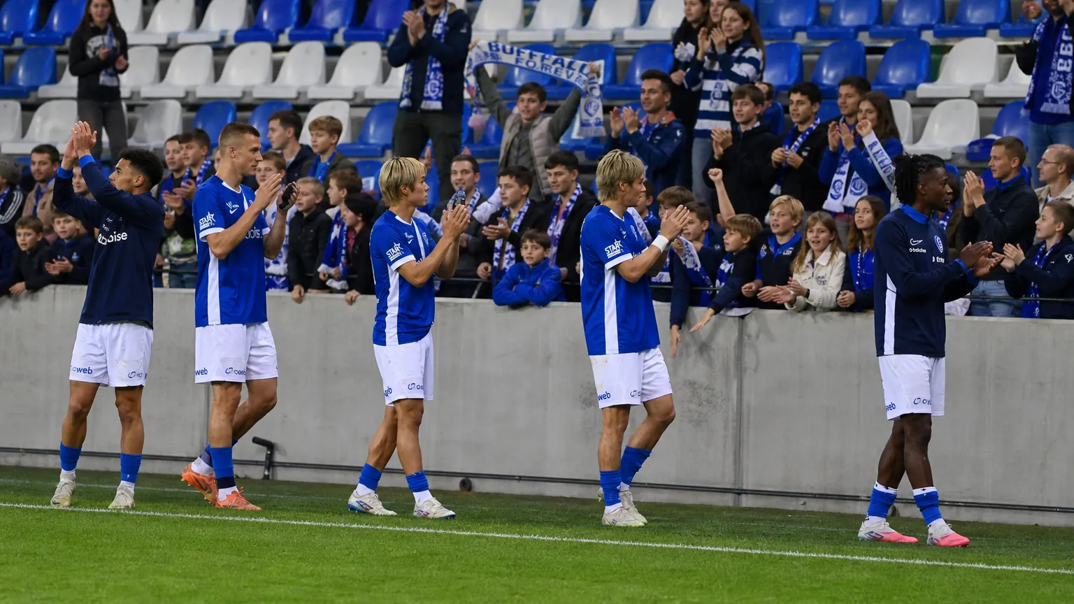 Voetballers in blauw-witte shirts groeten supporters langs de rand van het veld.