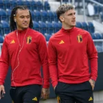 Twee voetballers in rode Belgische trainingstoppen lopen langs de tribunes van een stadion.