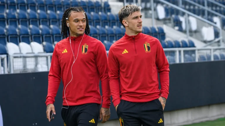 Twee voetballers in rode Belgische trainingstoppen lopen langs de tribunes van een stadion.
