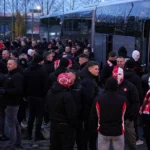 Een groep Antwerp-supporters verzamelt zich met rood-witte supportersattributen naast geparkeerde bussen.