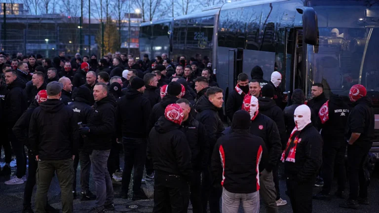 Een groep Antwerp-supporters verzamelt zich met rood-witte supportersattributen naast geparkeerde bussen.