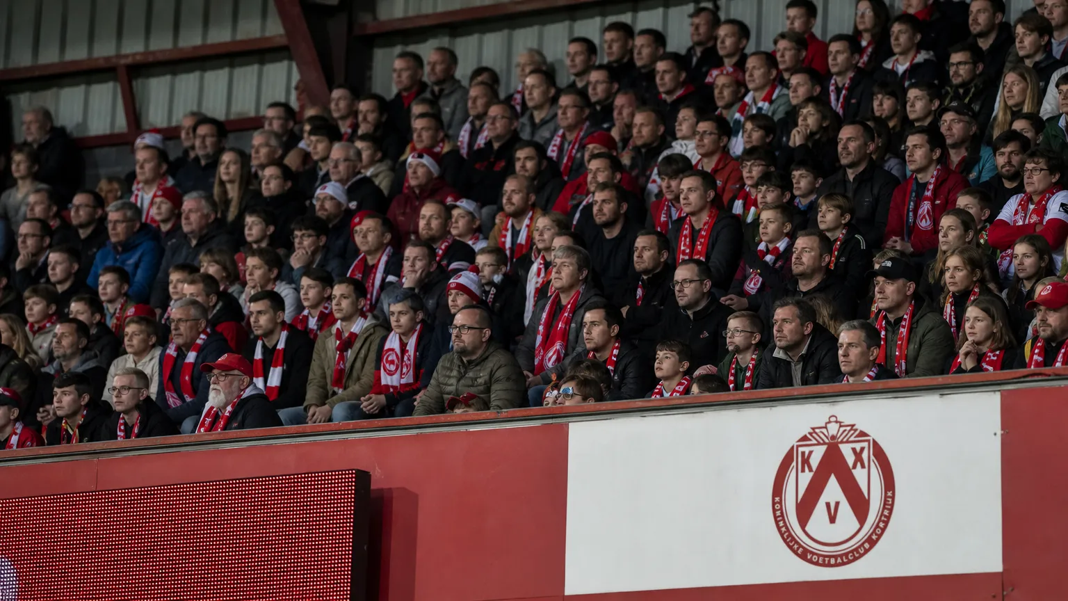 Een groep supporters van KV Kortrijk op de thuistribune, met een clubembleem op een tribune­bord.
