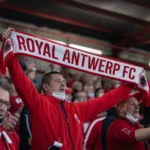 Antwerp-supporters houden een rood-witte supporterssjaal omhoog in een overdekte tribunezone.