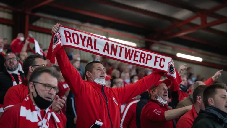 Antwerp-supporters houden een rood-witte supporterssjaal omhoog in een overdekte tribunezone.