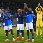 Een groep KRC Genk-spelers applaudisseert naar de thuistribune onder stadionlichten.