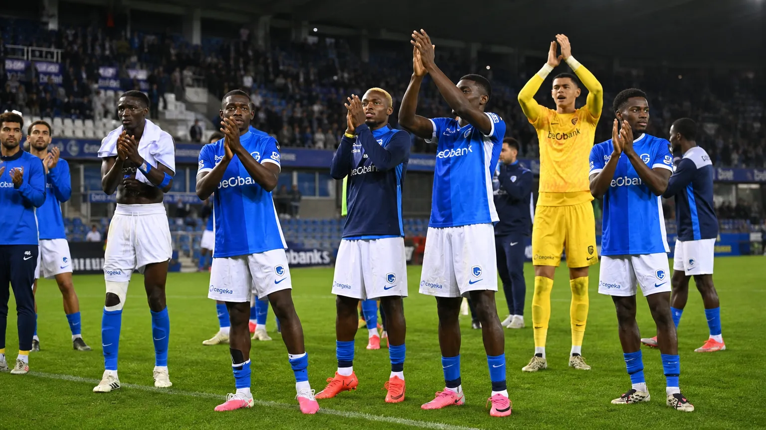 Een groep KRC Genk-spelers applaudisseert naar de thuistribune onder stadionlichten.