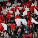 Een volle groep supporters zwaait met rode en witte vlaggen op de tribune.