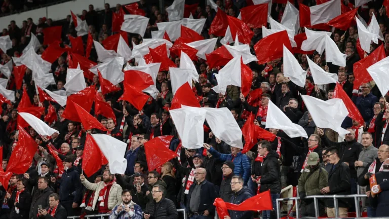Een volle groep supporters zwaait met rode en witte vlaggen op de tribune.