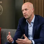 Jordi Cruijff spreekt in een Ajax-medialokaal met een Ajax-embleem op de wand.