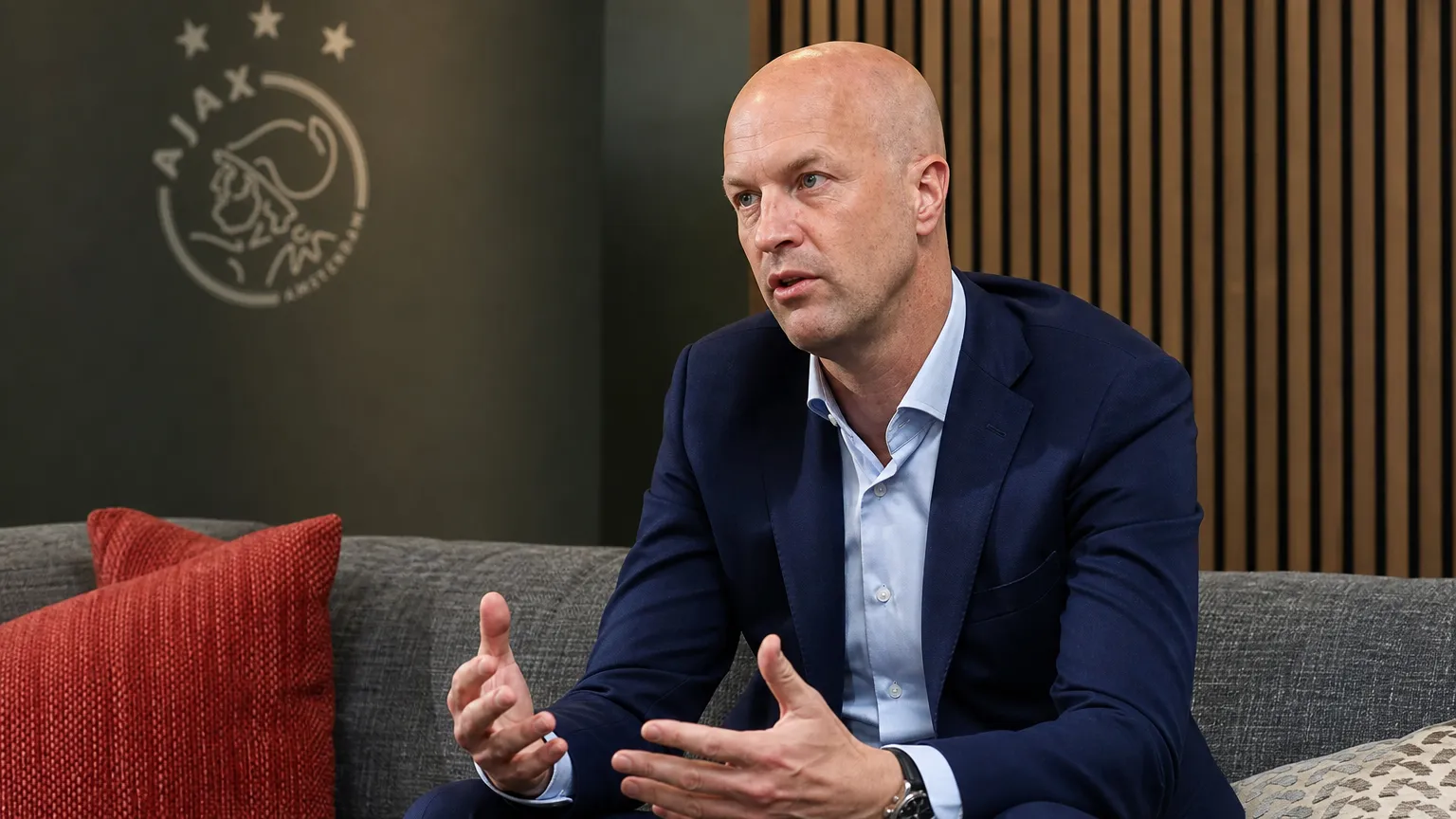 Jordi Cruijff spreekt in een Ajax-medialokaal met een Ajax-embleem op de wand.