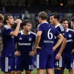 Een groep voetballers in paarse tenues overlegt op het veld na een wedstrijd.