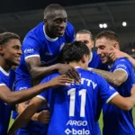 Een groep voetballers in blauwe wedstrijdtenues viert samen op het veld.
