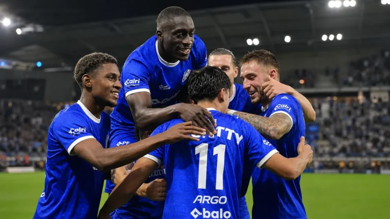 Een groep voetballers in blauwe wedstrijdtenues viert samen op het veld.