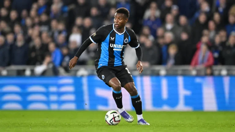 Een voetballer in een donker tenue met blauwe strook controleert de bal in een avondstadion.