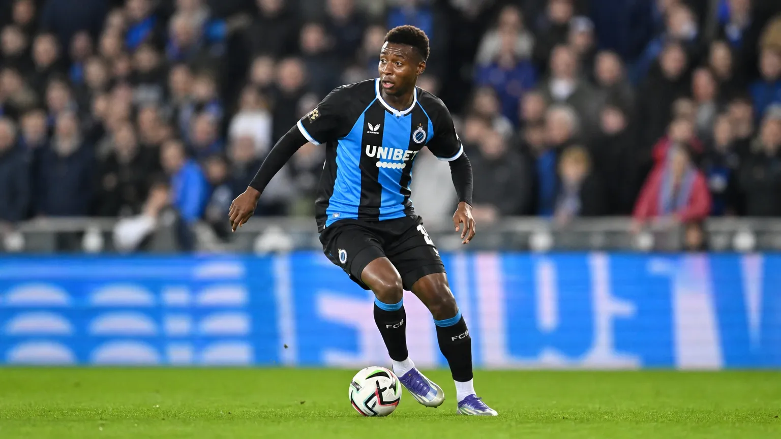Een voetballer in een donker tenue met blauwe strook controleert de bal in een avondstadion.