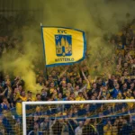 Supporters van KVC Westerlo met geel-blauwe vlaggen en rook achter het doel.