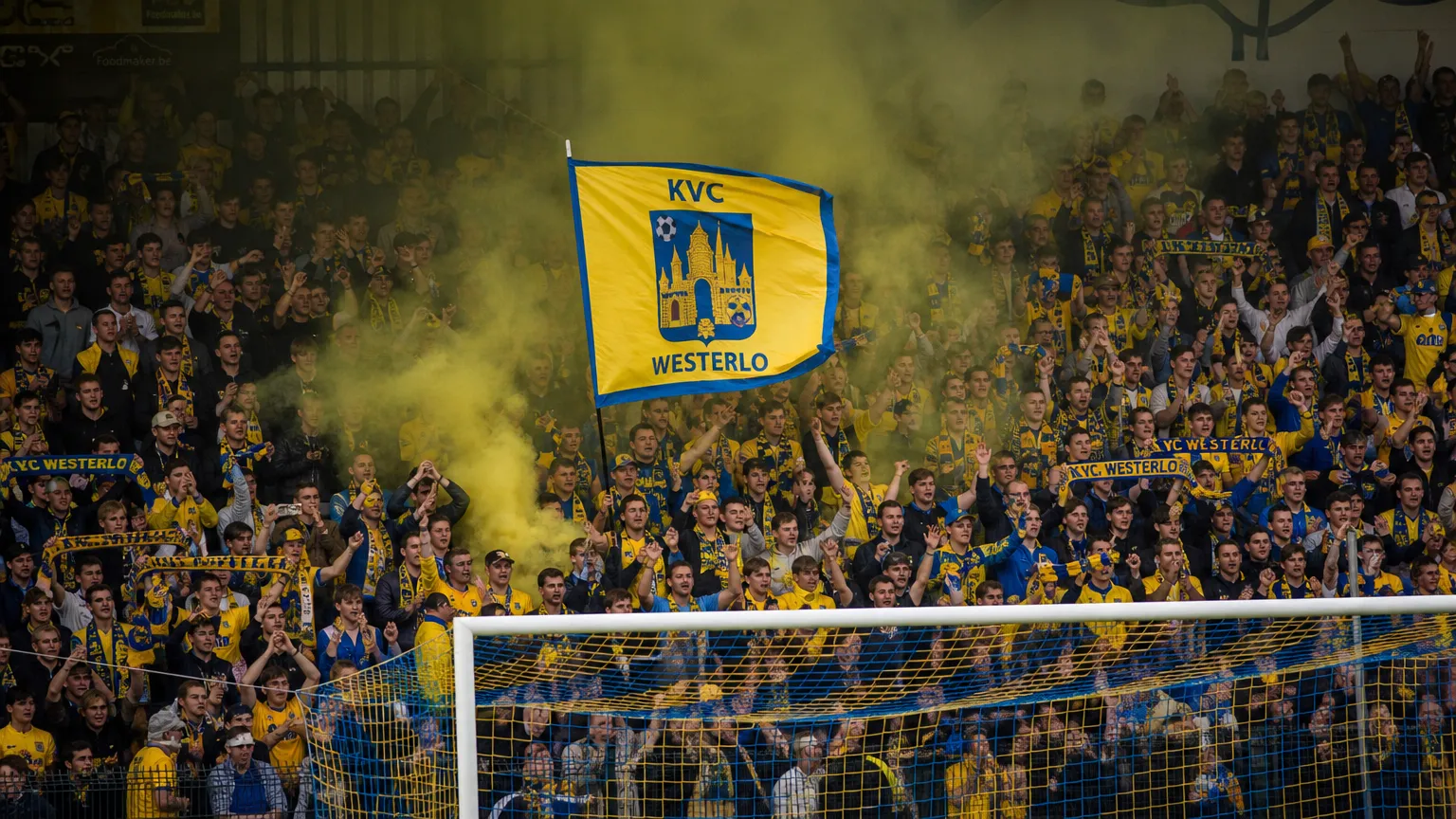 Supporters van KVC Westerlo met geel-blauwe vlaggen en rook achter het doel.