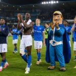 Een groep voetballers in blauwe shirts en een mascotte applaudisseren op een verlicht veld.
