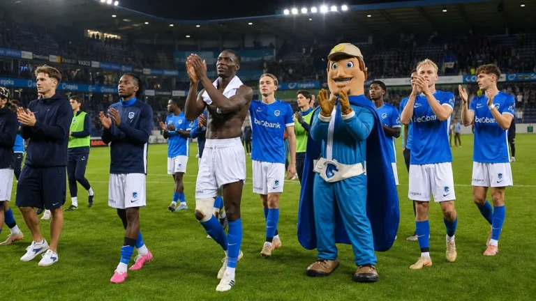 Een groep voetballers in blauwe shirts en een mascotte applaudisseren op een verlicht veld.