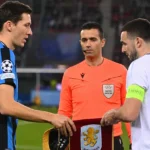 Een speler van Club Brugge en een speler van Aston Villa bij een Europese wedstrijd, met een scheidsrechter ertussen.