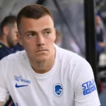 Een KRC Genk-speler in een wit wedstrijdshirt zit aandachtig in de dug-out.
