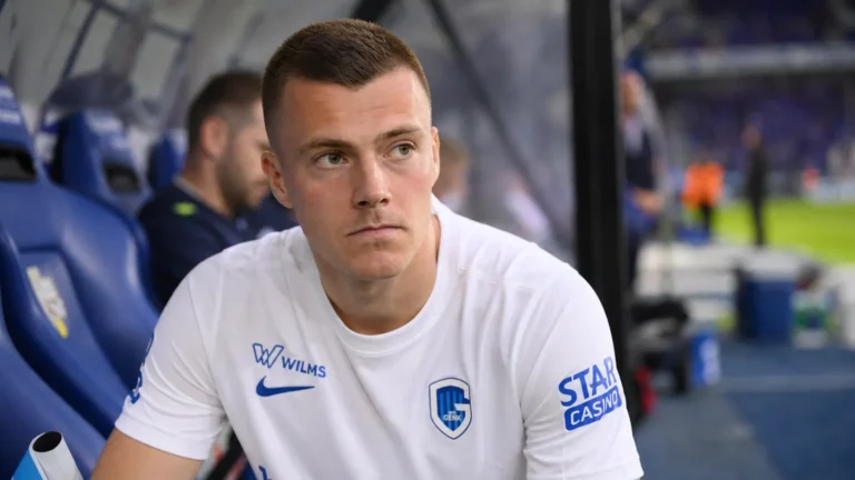 Een KRC Genk-speler in een wit wedstrijdshirt zit aandachtig in de dug-out.