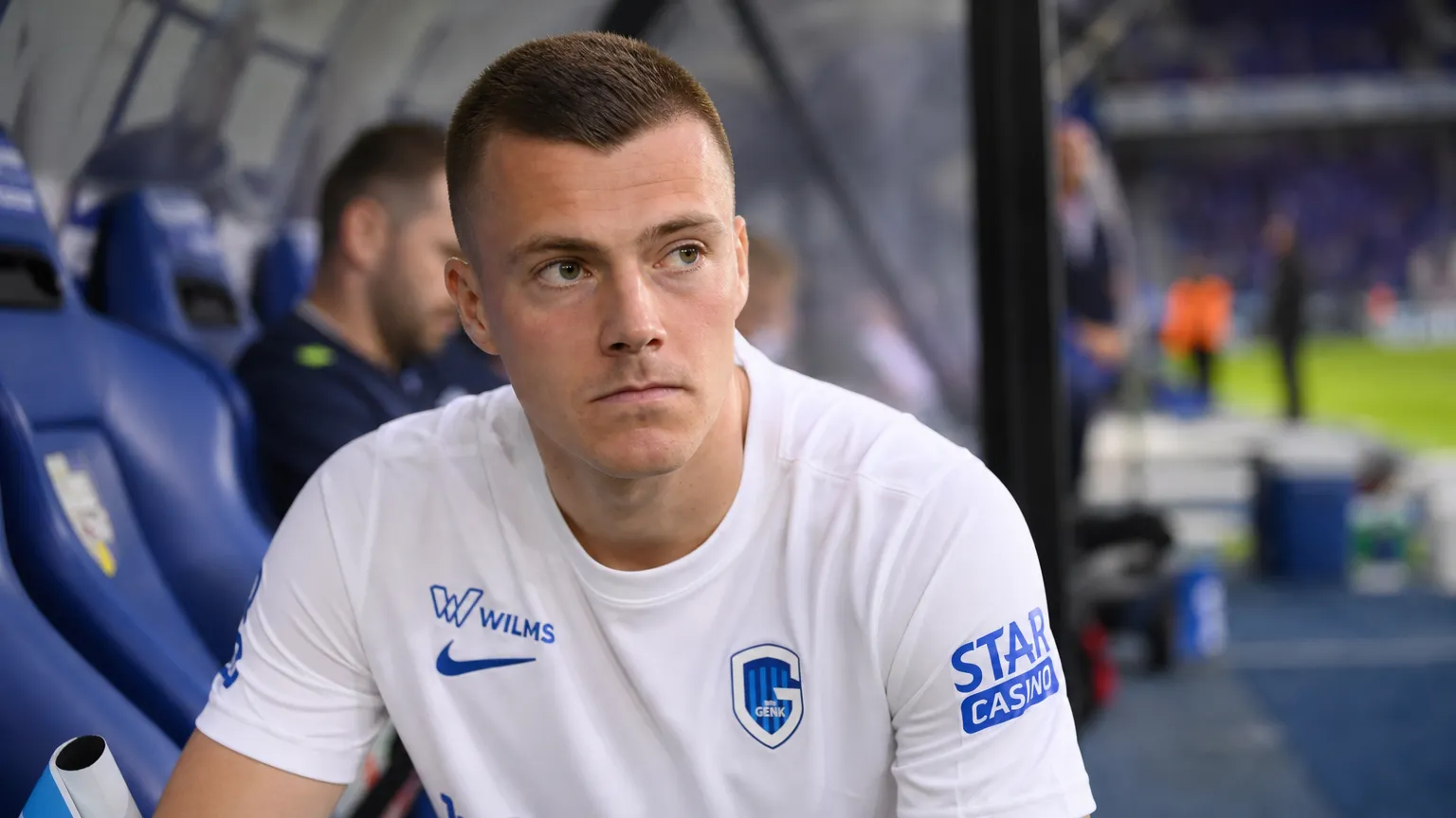 Een KRC Genk-speler in een wit wedstrijdshirt zit aandachtig in de dug-out.